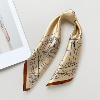 Pale apricot silk scarf with black line art depicting cityscape architecture, folded to display shimmering fabric and brown border, on neutral background.