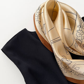 Pale apricot silk bandana scarf with elegant black line art design and cognac border, displayed alongside a black fabric on white background