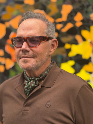 Styling example: brown men's silk scarf with green horseshoe pattern worn with brown polo shirt and amber sunglasses in outdoor setting