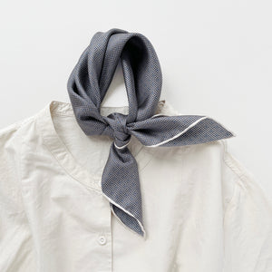 a classic silk scarf neckerchief featuring black and white checks and navy blue polka dots with white hand-rolled hems, knotted as a neckerchief, paired with a light beige shirt  Edit alt text