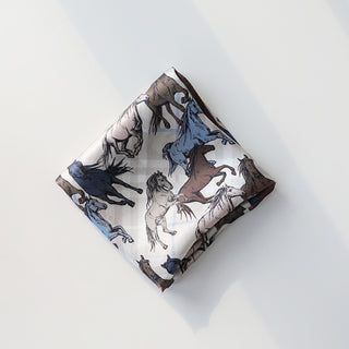 Luxury silk scarf with wild horse pattern featuring galloping horses in brown, blue, and cream colors, folded into a square on white background