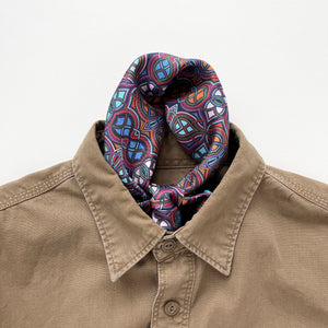 a silk men's neckerchief scarf featuring an abstract intricate pattern in plum purple, blue and orange hues, knotted and tucked into a men's khaki shirt
