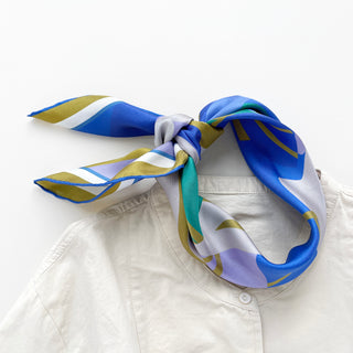Blue abstract lotus silk scarf tied around collar of beige shirt, featuring vibrant blue, lavender, teal, and mustard yellow geometric patterns