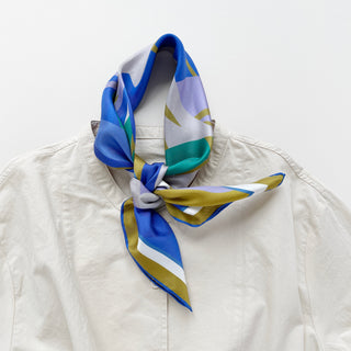 Abstract lotus silk scarf in vibrant blue, green, lilac, and yellow patterns tied as a neckerchief on a beige jacket collar