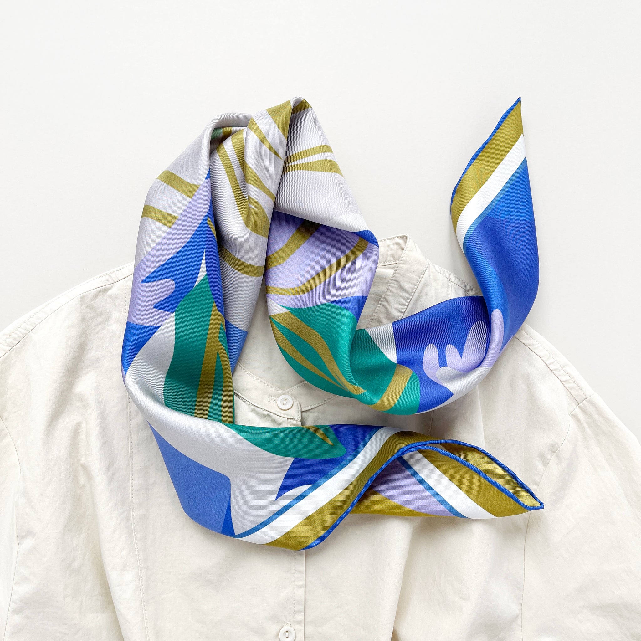 an abstract lotus print square silk scarf in blue, green, lilac and mustard yellow palette with hand-rolled hems paired with a light beige women's turtle neck shirt