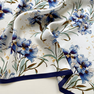 Close-up of women's silk scarf with delicate blue bell flowers pattern on white background with green stems and subtle gold accents.