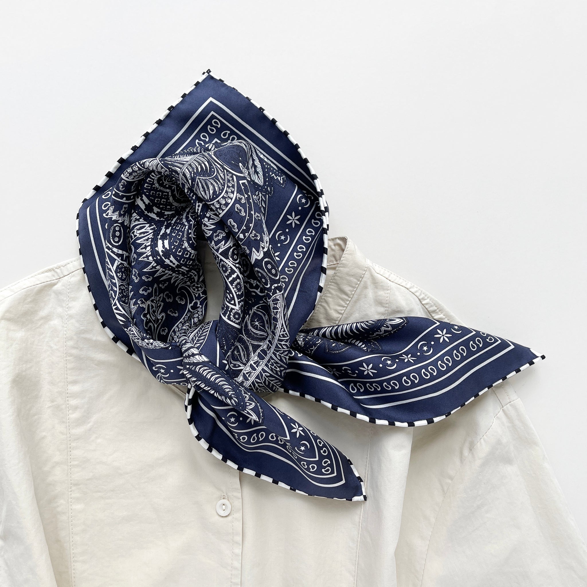 a navy blue silk bandana scarf featuring white and black symmetric pattern with striped hand-rolled edges, knotted as a neck scarf, paired with a light beige turtle neck shirt