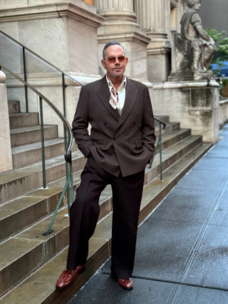 Man wearing a beige and red horseshoe print silk scarf with a dark brown double-breasted suit, a white shirt, sunglasses, and brown shoes stands on stone steps in front of a classical building.