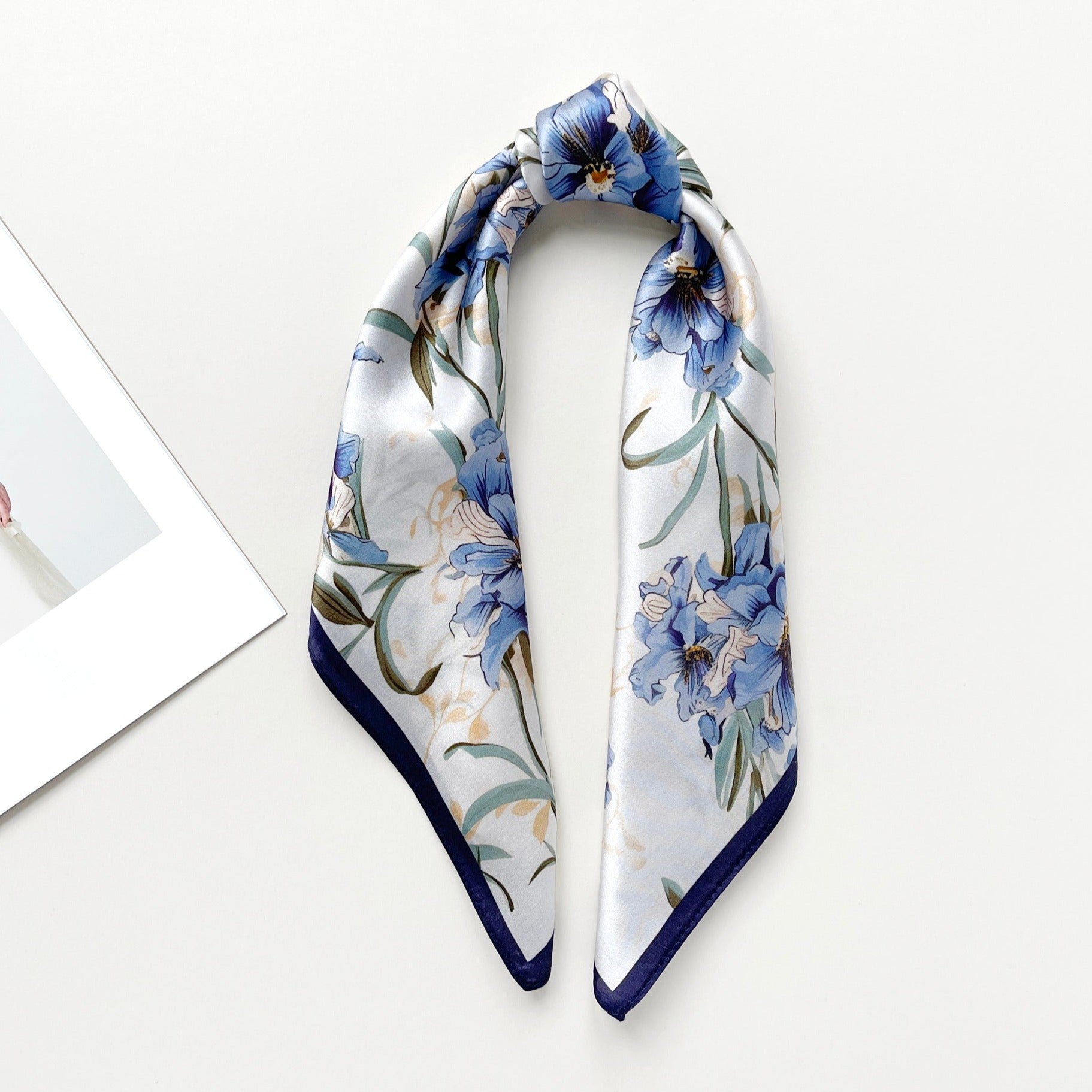 a silk bandana scarf featuring blue bell print with a white base and navy blue edges tied as a ponytail head scarf