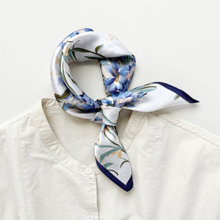Elegant silk bandana scarf with blue bell floral pattern on white background, tied around collar of cream blouse with navy blue border trim