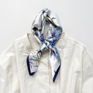 Square silk bandana scarf with blue bell floral pattern on white background, elegantly tied at neck of cream blouse with navy trim