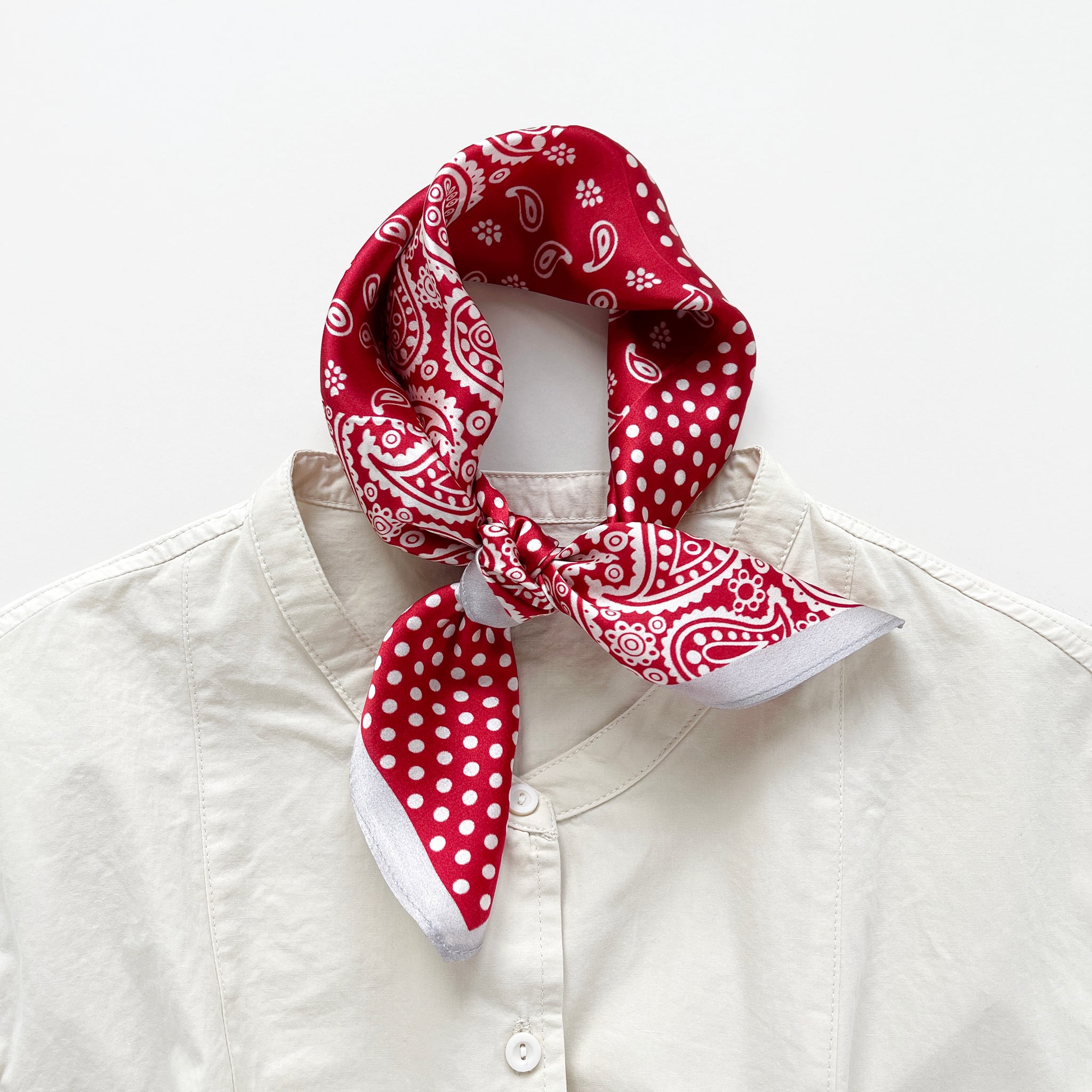 a vibrant red silk bandana scarf featuring paisley and polka dot pattern, knotted as a neckerchief, paired with a light beige turtle neck shirt