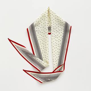 A folded silk scarf with a cream base, featuring a delicate black cross pattern, detailed black borders, and a red border trim, displayed on a light gray background.