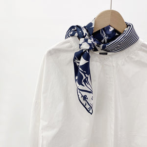 a navy blue and white square silk scarf featuring the combination of floral motifs, polka dots, and stripes prints with hand-rolled edges knotted as a neck scarf, paired with a women's white shirt
