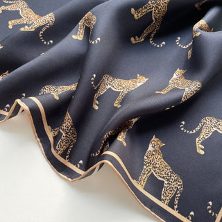 Luxurious black silk scarf with gold leopard pattern, featuring hand-rolled beige border. Smooth silk twill texture elegantly draped showing the fabric's fluidity.