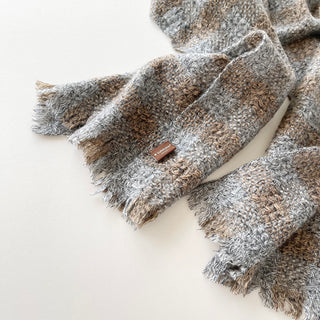 A close-up of a soft, textured knit cashmere scarf  in a heather mix of grey, beige, and brown tones, with fringed ends and LuluLane® label.