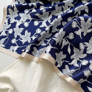 Navy blue silk bandana scarf with elegant white floral pattern and light beige border trim, showcasing luxurious fabric texture