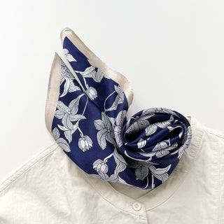 Square silk scarf with navy blue background and white botanical leaf pattern, elegantly styled around collar of cream blouse