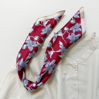 Red silk bandana with blue botanical leaf pattern styled as neckerchief on white blouse, showing luxury fabric texture and floral design