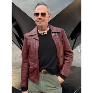 Man wearing a beige and red horseshoe print silk scarf with a burgundy leather jacket, black sweater and sunglasses.