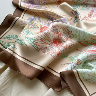 Elegant beige silk scarf with delicate floral pattern in coral, blue and green, featuring a rich brown border, displayed in soft folds