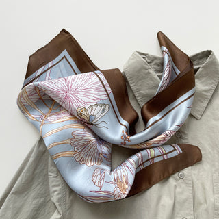 Light blue silk scarf with delicate floral pattern and rich brown border, elegantly draped over light beige shirt, showcasing versatile styling options.
