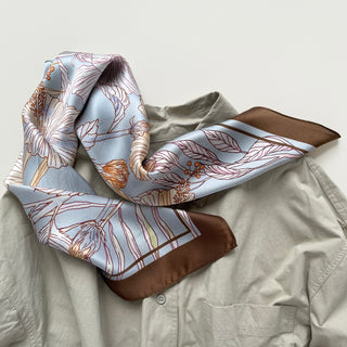 Light blue silk bandana scarf with delicate floral outline print in rust and pink tones, featuring brown border, draped on beige shirt