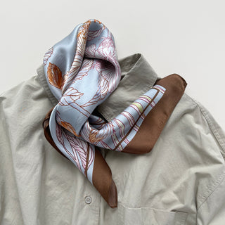 Light blue silk bandana scarf with delicate floral outline pattern in pink and orange, featuring brown border, elegantly draped on beige shirt collar