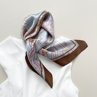Light blue silk scarf with delicate floral line art in pink and orange, featuring a rich brown border, elegantly draped over a white garment.