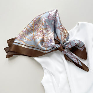 Light blue silk scarf with delicate floral outline pattern in burgundy and gold, featuring a rich brown border, elegantly knotted on white fabric.