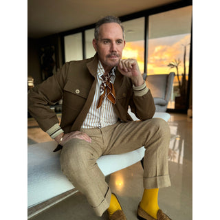 Man wearing a brown tones silk square neckerchief featuring retro geometric pattern in a stylish outfit sitting in a modern room with a sunset view.