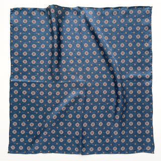 Denim blue silk neckerchief with repeating taupe medallion pattern, displayed flat showing hand-rolled edges and luxurious twill texture
