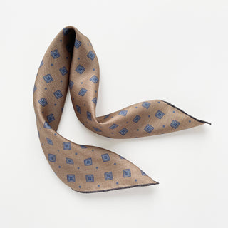 Luxury taupe silk neckerchief with blue diamond medallion pattern, hand-rolled edges, displayed in an elegant S-curve on white background