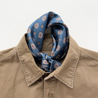 Denim blue silk neckerchief with medallion pattern styled on taupe shirt collar, showing elegant knot and luxurious fabric texture