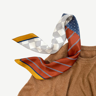Navy and rust silk neckerchief with geometric patchwork pattern featuring white checkered design, blue medallions, and rust stripes, draped on camel sweater