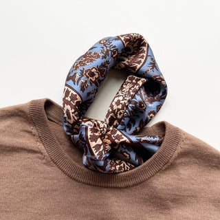 Blue silk neckerchief with brown paisley pattern elegantly tied around a light brown sweater collar, showcasing luxurious fabric texture.