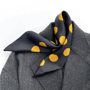 a black silk scarf with hand-rolled hems and golden polka dots laying on a dark grey wool coat