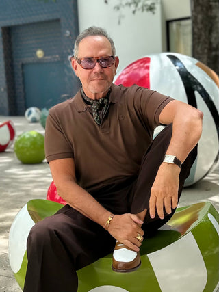A man wearing a brown and green horseshoe print silk scarf with a brown polo shirt, brown trousers, and sunglasses sits on a green and white mushroom-shaped bench in an outdoor setting.