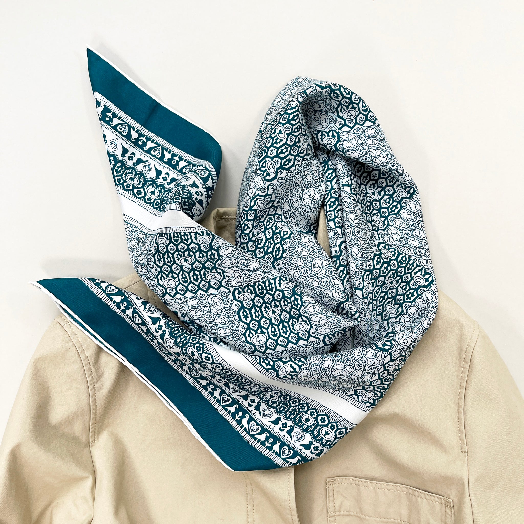 a turquoise blue square silk scarf with birds, flowers and trees prints laying on a beige coat