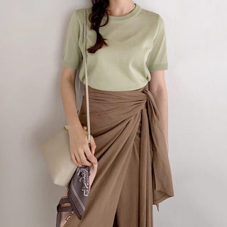 Person wearing sage green t-shirt and brown wrap skirt, holding a patterned bandana with cream shoulder bag, demonstrating how to style the accessory.