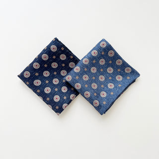 Two luxury silk neckerchiefs with circular medallion pattern, displayed side by side - one is denim blue and the other is dark denim blue, both with hand-rolled edges.