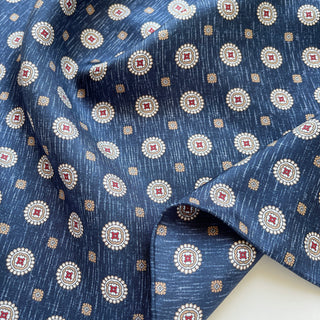 Dark denim blue silk neckerchief with elegant medallion pattern featuring cream circles, burgundy centers, and gold accents, draped to show luxurious texture