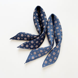 Two luxury silk neckerchiefs with circular medallion pattern, displayed side by side - one is denim blue and the other is dark denim blue, both with hand-rolled edges.