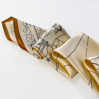 Pale apricot silk scarf with black line art cityscape design and caramel-colored border, displaying a soft sheen and elegant drape
