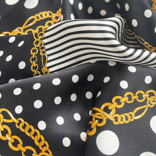 Luxury silk scarf with black background featuring white polka dots, black and white stripes, and decorative gold chain pattern, showing glossy satin finish