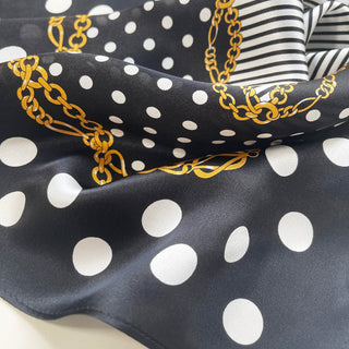 Luxurious black silk scarf with white polka dots, gold chain pattern, and striped border, showcasing glossy satin texture and elegant draping.