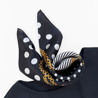 Black silk scarf with white polka dots, gold chain print, and striped pattern, elegantly styled in a twisted knot against white background