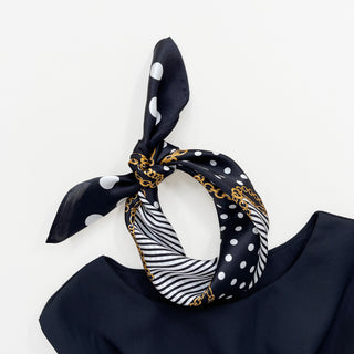 Black silk scarf with white polka dots, golden chain patterns, and black and white stripes, tied in a stylish knot on dark fabric.