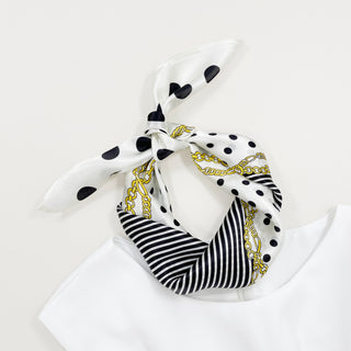 White silk scarf with black polka dots, stripes and gold chain pattern, elegantly tied in a bandana style on white background