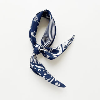 Navy blue and white patterned silk scarf featuring mixed floral, polka dot, and animal print motifs on a light grey board